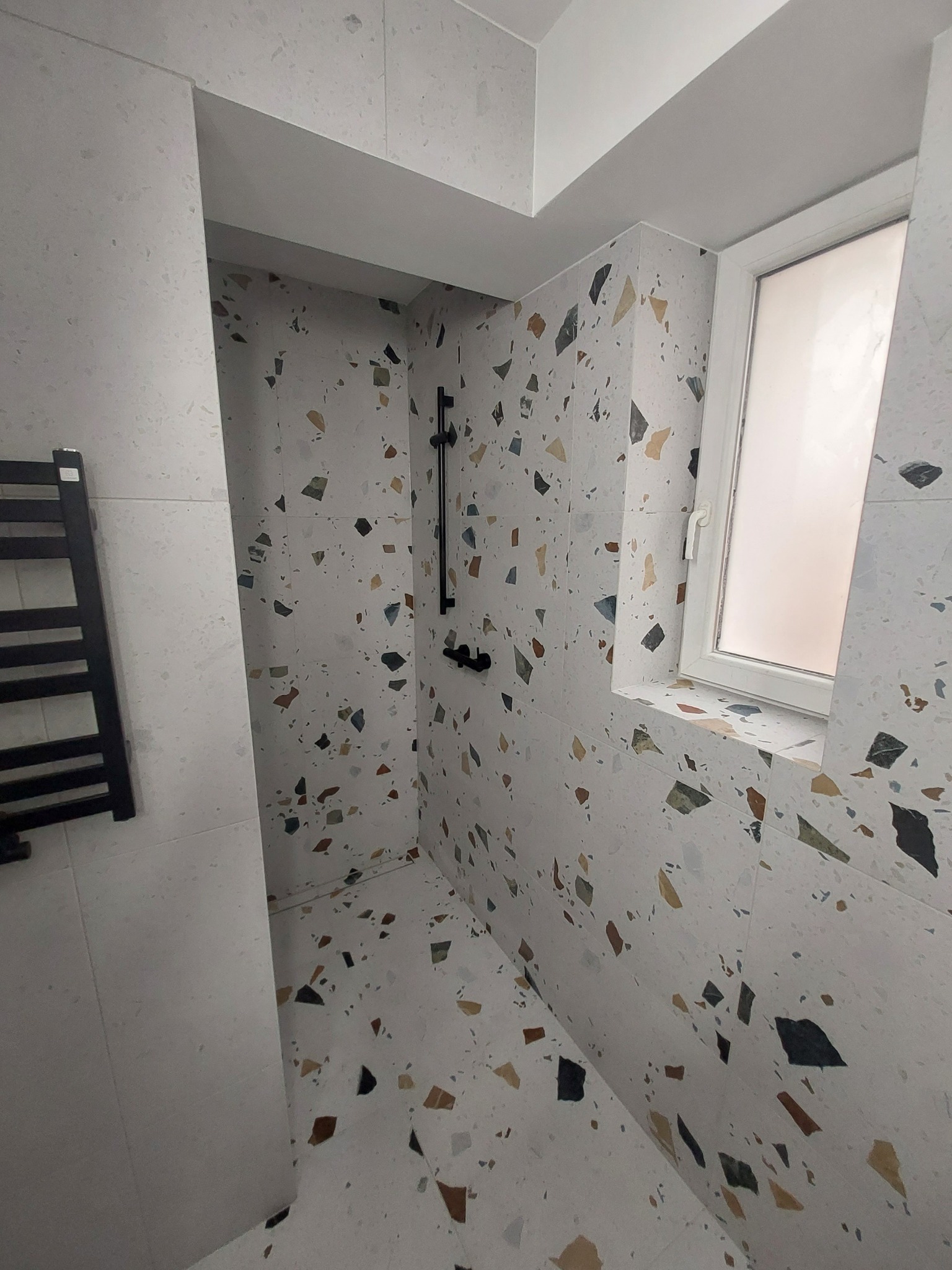 Wykończona łazienka z kabiną prysznicową wyłożona płytkami w stylu lastryko, z czarną armaturą i grzejnikiem drabinkowym.