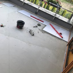 Kompleksowe wykończenia wnętrz i zewnętrzne - Montaż płytek na balkonie z widocznymi narzędziami, poziomnicami, wiadrem z zaprawą oraz kwiatami w donicach na balustradzie w tle.