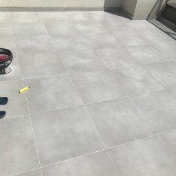 Kompleksowe wykończenia wnętrz i zewnętrzne - Jasnoszare płytki ceramiczne na balkonie, wiadro z farbą i nóż tapicerski w lewym rogu, częściowo widoczne buty