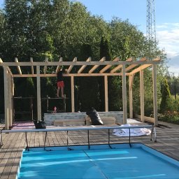 Kompleksowe wykończenia wnętrz i zewnętrzne - Budowa drewnianej pergoli przy basenie: mężczyzna na rusztowaniu montuje konstrukcję, widoczne materiały budowlane i narzędzia, w tle zieleń ogrodu i maszt telekomunikacyjny.