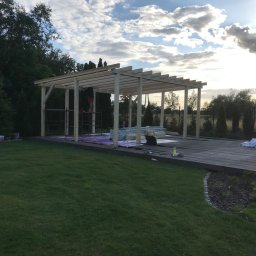 Kompleksowe wykończenia wnętrz i zewnętrzne - Konstrukcja drewnianej pergoli w trakcie montażu na tarasie, widok na zielony trawnik i zachmurzone niebo.