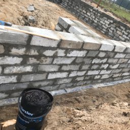 Kompleksowe wykończenia wnętrz i zewnętrzne - Wznoszenie muru fundamentowego z bloczków betonowych, widoczny kubeł z preparatem hydroizolacyjnym Dysperbit i narzędzia murarskie na górnej krawędzi muru.