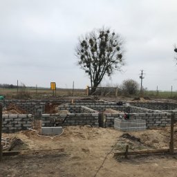 Kompleksowe wykończenia wnętrz i zewnętrzne - Budowa fundamentów z bloczków betonowych w Łomiankach na tle pochmurnego nieba i drzew z jemiołą.