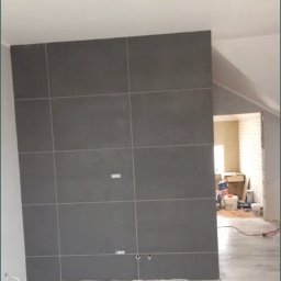 Kompleksowe wykończenia wnętrz i zewnętrzne - Ściana wyłożona szarymi płytkami wielkoformatowymi, z widocznymi otworami na instalacje, na tle podłogi z paneli imitujących drewno, w trakcie prac wykończeniowych.