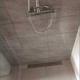 Kompleksowe wykończenia wnętrz i zewnętrzne - Szare płytki ceramiczne na ścianach i suficie kabiny prysznicowej z odpływem liniowym i chromowaną deszczownicą.