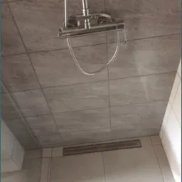 Szare płytki ceramiczne na ścianach i suficie kabiny prysznicowej z odpływem liniowym i chromowaną deszczownicą.