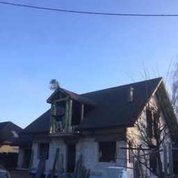 Kompleksowe wykończenia wnętrz i zewnętrzne - Dom w stanie surowym zamkniętym z widoczną konstrukcją dachu i oknami, z osobą stojącą w otworze okiennym na poddaszu, widok z zewnątrz w słoneczny dzień.