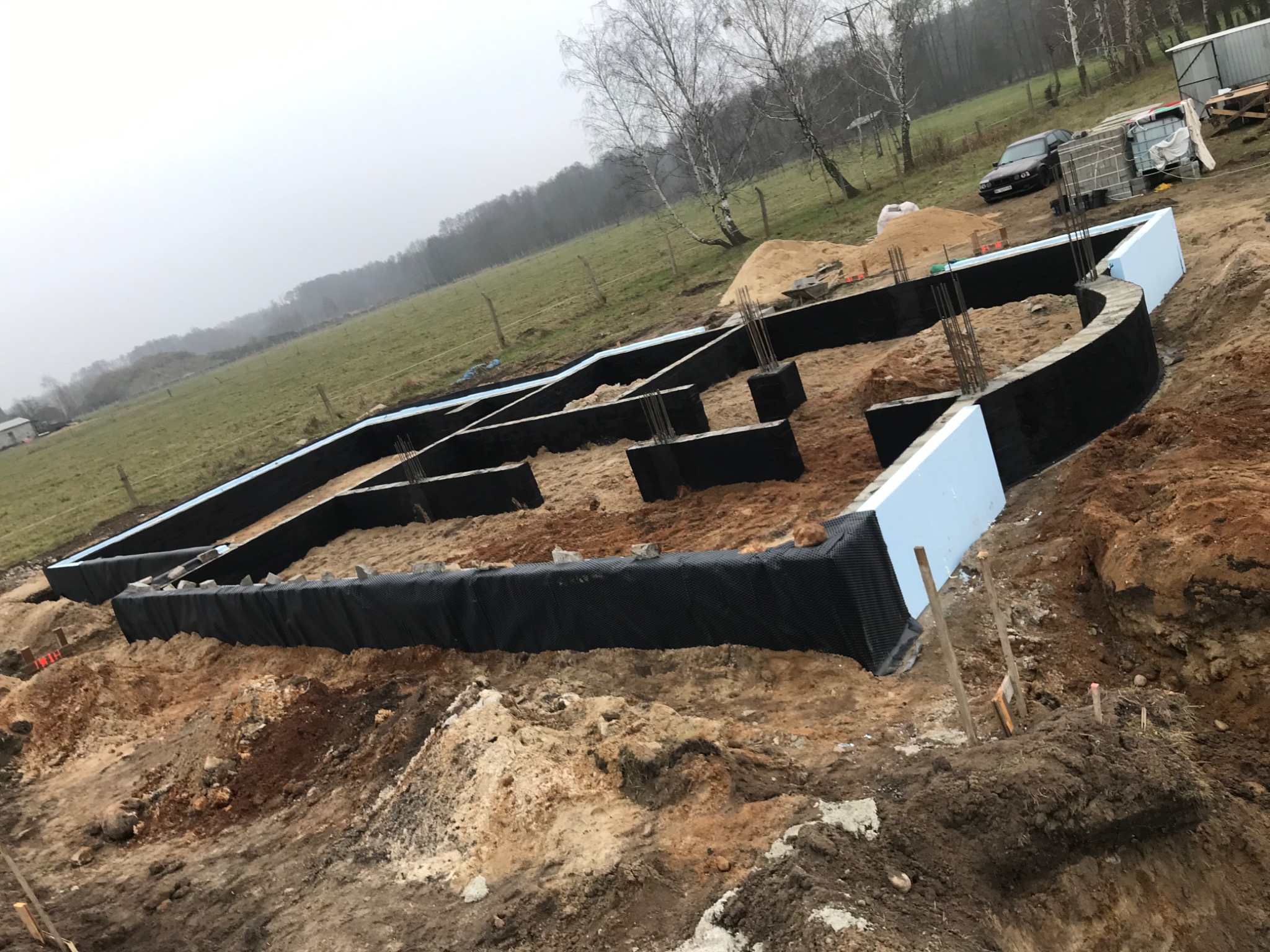 Przygotowany fundament z izolacją pionową i termiczną, widoczne zbrojenie, teren budowy w trakcie prac ziemnych, w tle łąka i las.