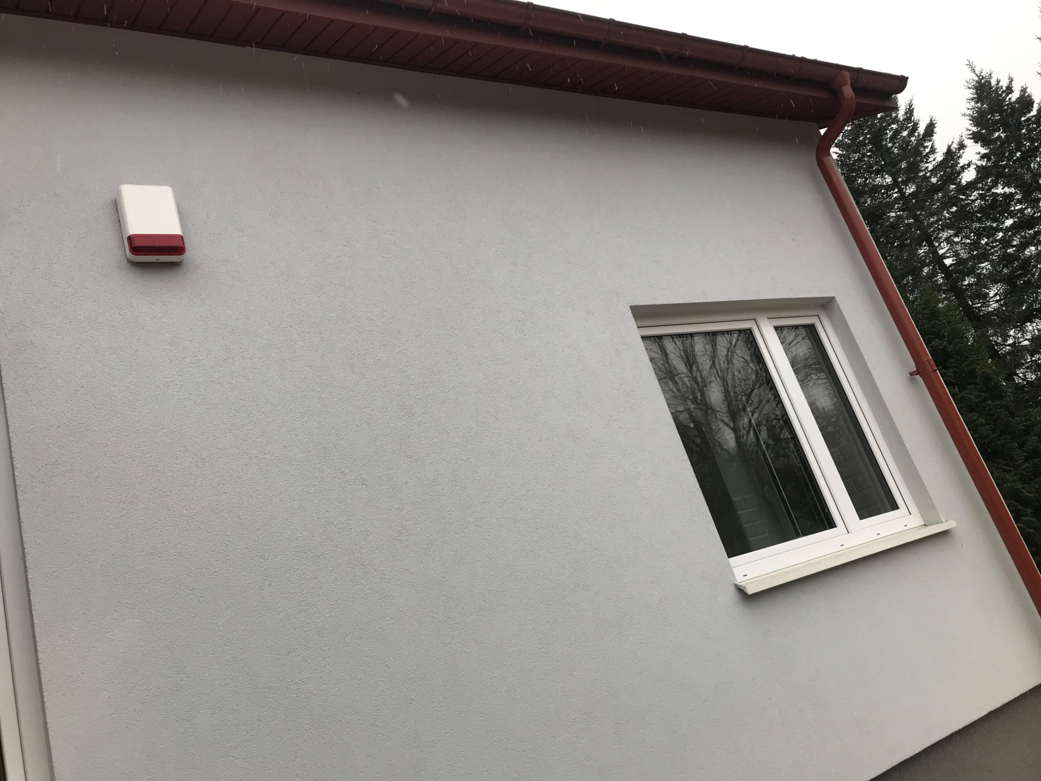 Jasnoszara elewacja budynku z oknem, brązowym orynnowaniem i alarmem, widoczne drzewa w tle.
