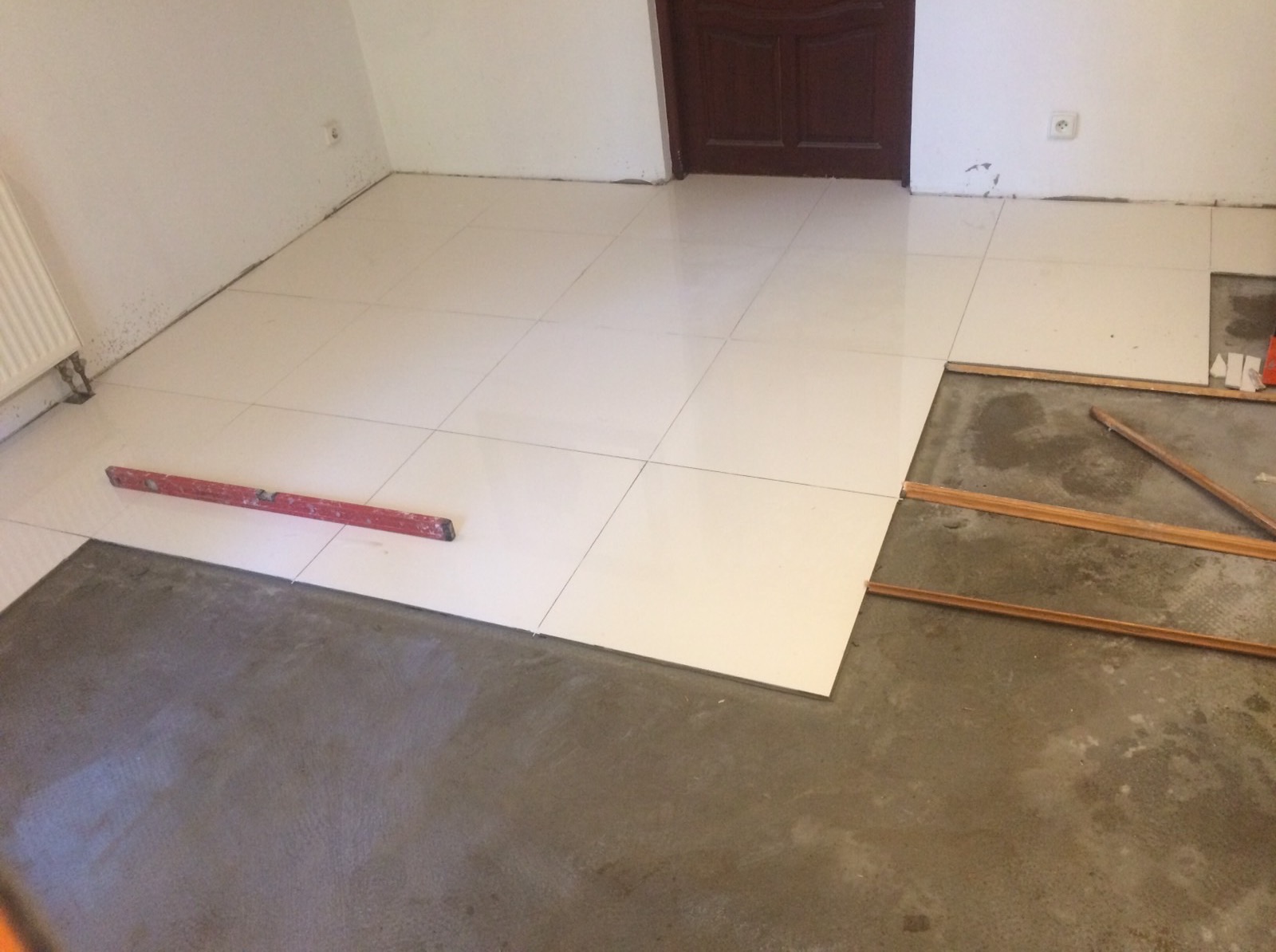 Układanie białych, kwadratowych płytek podłogowych w pomieszczeniu z szarą wylewką, widoczna poziomica i fragmenty listew przypodłogowych.