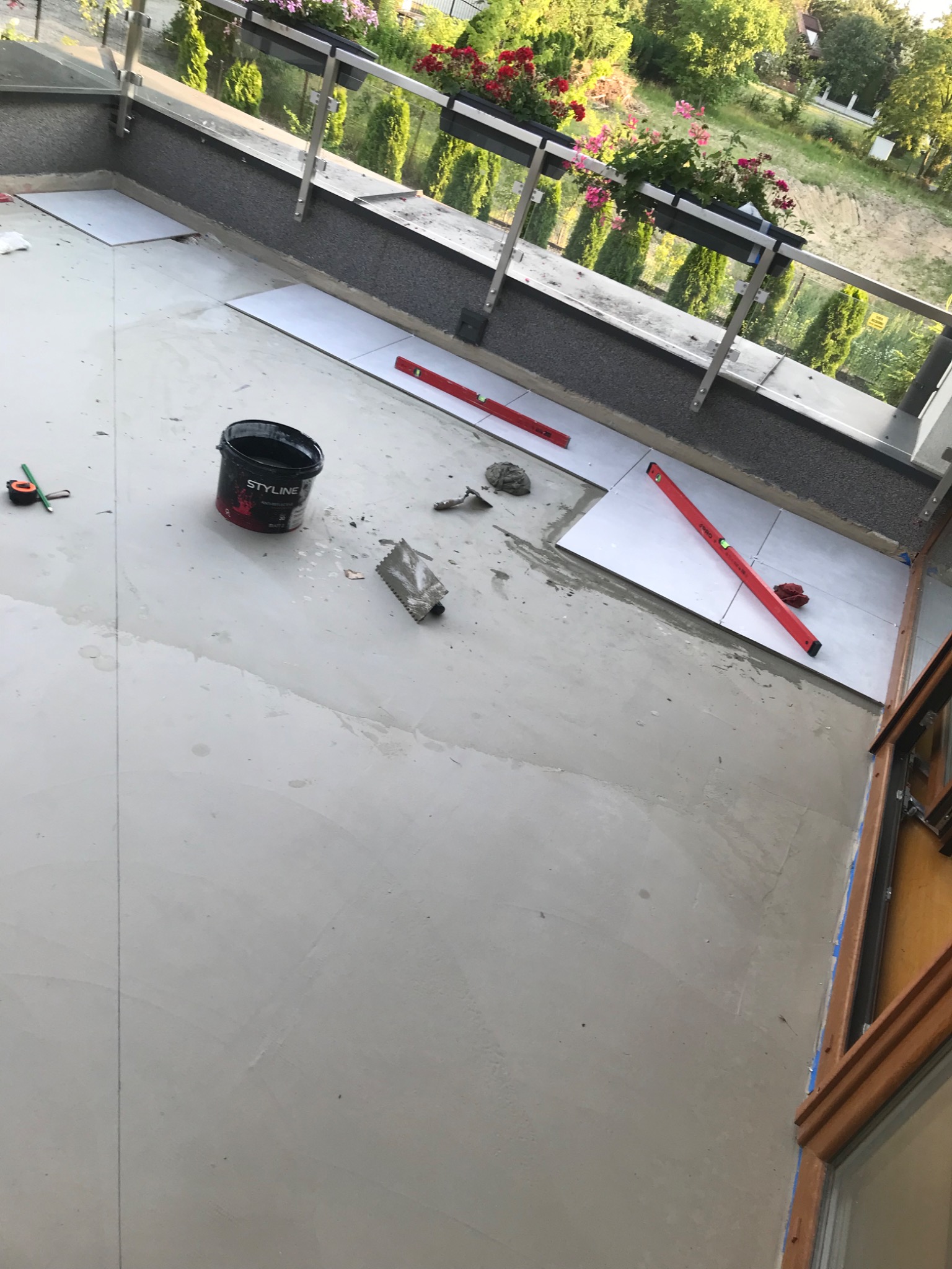 Montaż płytek na balkonie z widocznymi narzędziami, poziomnicami, wiadrem z zaprawą oraz kwiatami w donicach na balustradzie w tle.