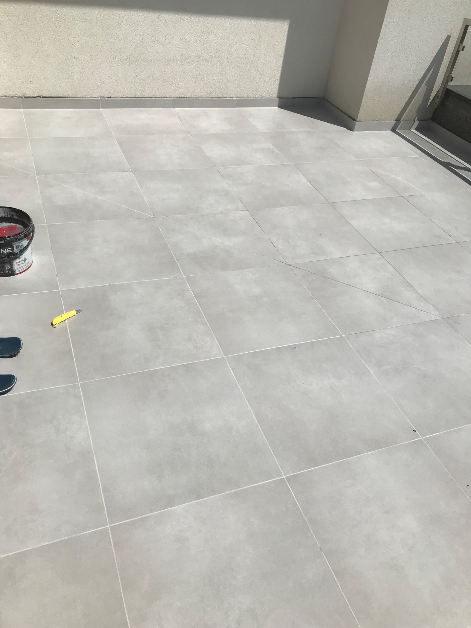 Jasnoszare płytki ceramiczne na balkonie, wiadro z farbą i nóż tapicerski w lewym rogu, częściowo widoczne buty