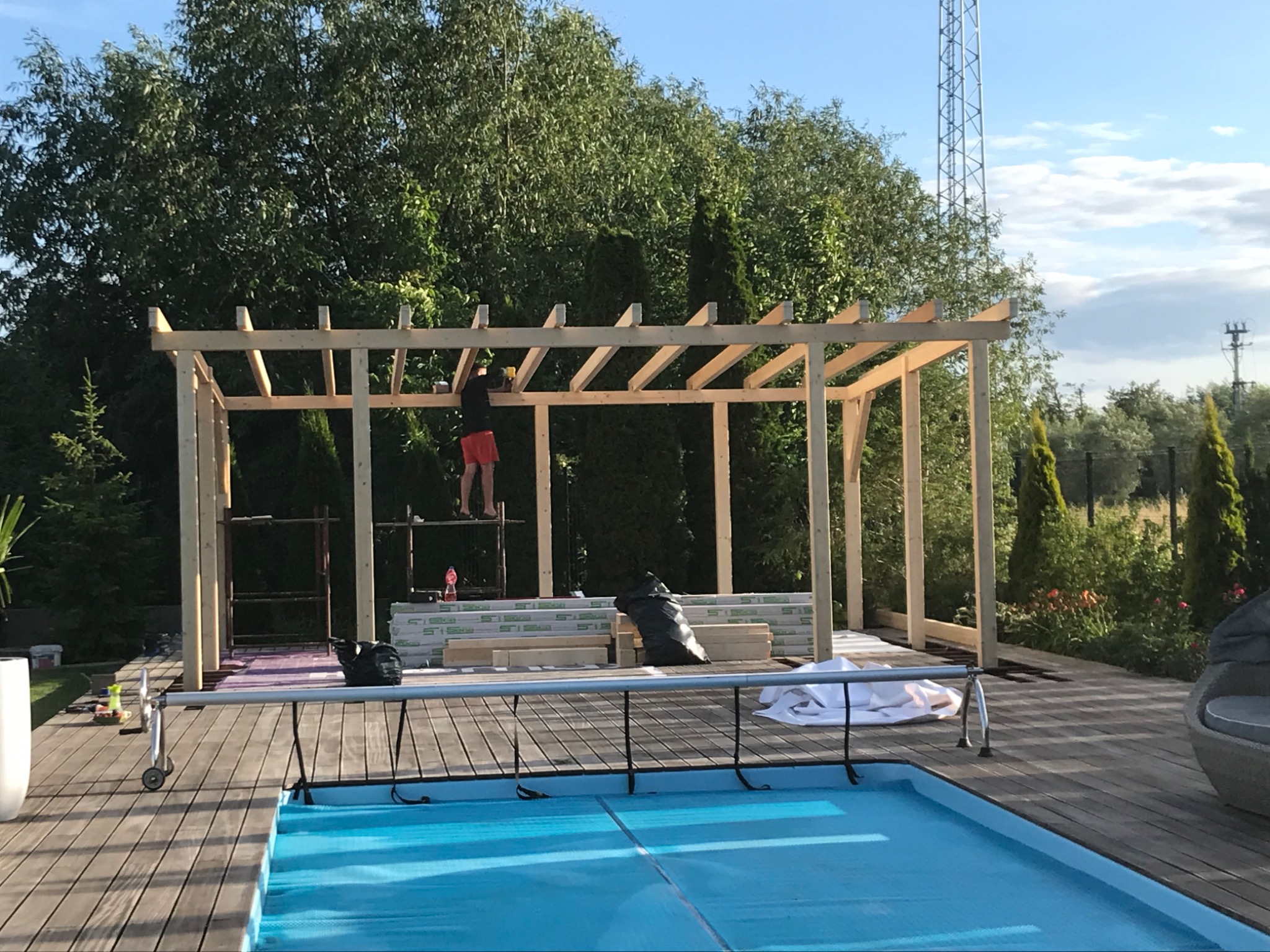 Budowa drewnianej pergoli przy basenie: mężczyzna na rusztowaniu montuje konstrukcję, widoczne materiały budowlane i narzędzia, w tle zieleń ogrodu i maszt telekomunikacyjny.