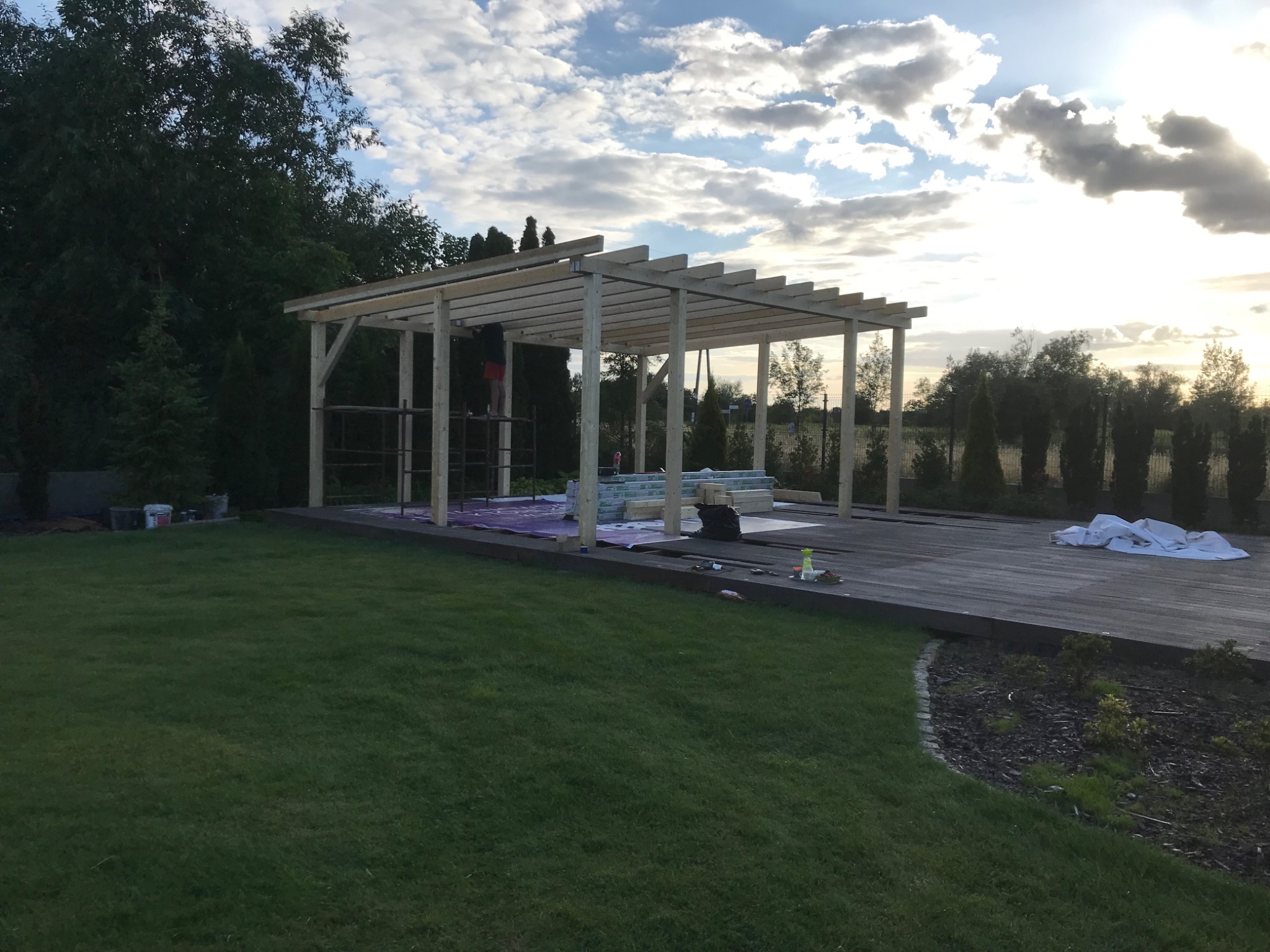 Konstrukcja drewnianej pergoli w trakcie montażu na tarasie, widok na zielony trawnik i zachmurzone niebo.