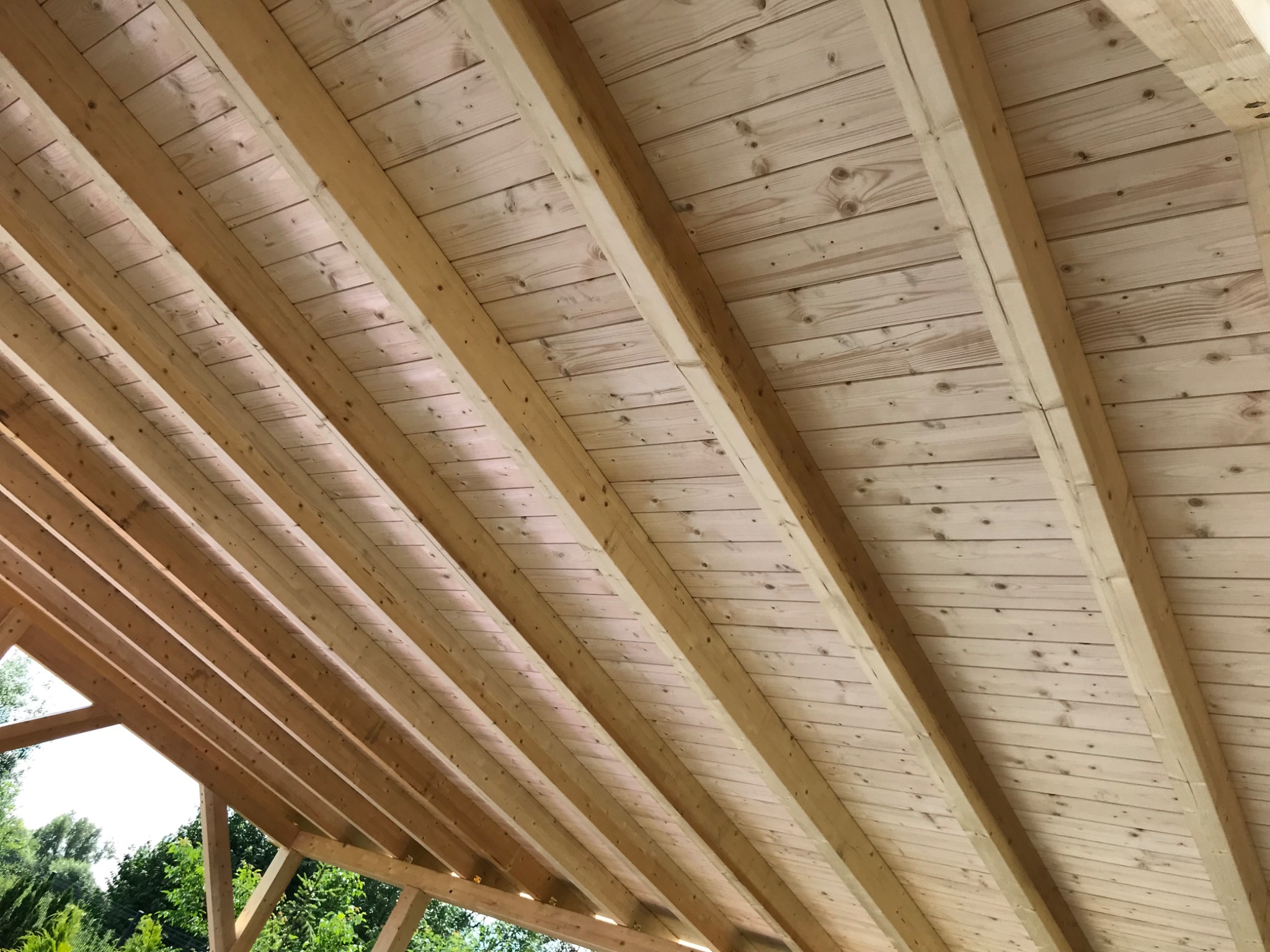 Surowy drewniany sufit z widocznymi belkami konstrukcyjnymi, widok z dołu na tle zieleni drzew.