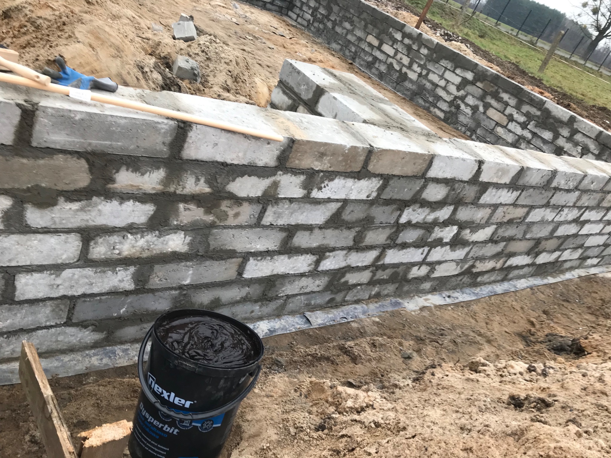 Wznoszenie muru fundamentowego z bloczków betonowych, widoczny kubeł z preparatem hydroizolacyjnym Dysperbit i narzędzia murarskie na górnej krawędzi muru.