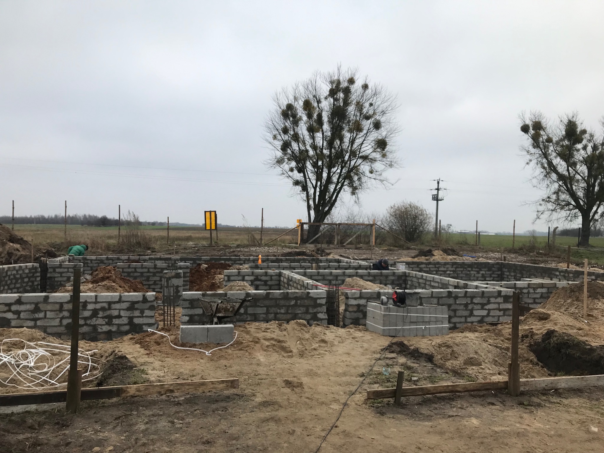 Budowa fundamentów z bloczków betonowych w Łomiankach na tle pochmurnego nieba i drzew z jemiołą.