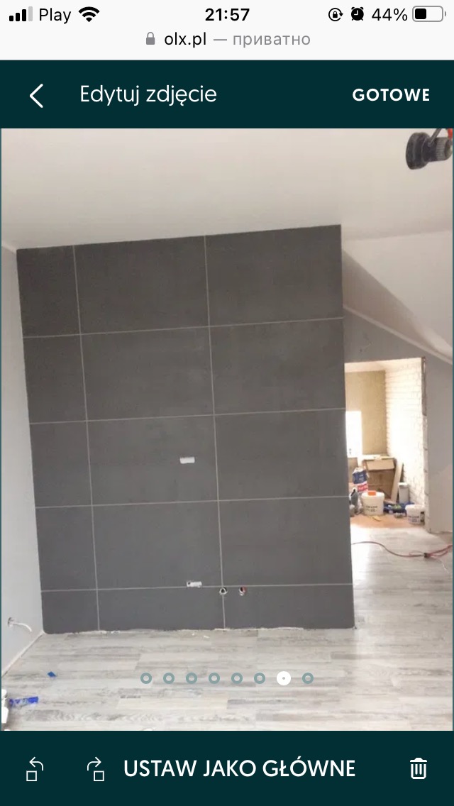 Ściana wyłożona szarymi płytkami wielkoformatowymi, z widocznymi otworami na instalacje, na tle podłogi z paneli imitujących drewno, w trakcie prac wykończeniowych.