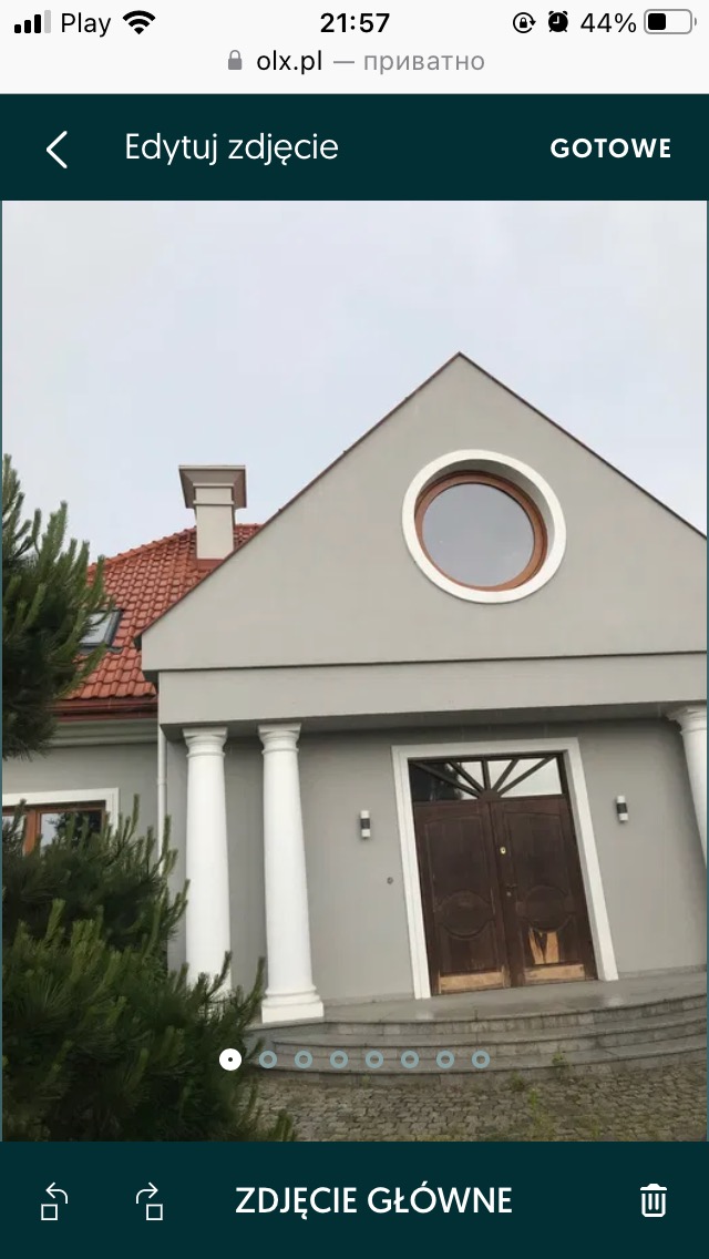 Szary dom jednorodzinny z kolumnami przy wejściu, okrągłym oknem na szczycie i czerwoną dachówką, widziany z dołu.