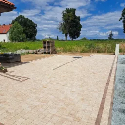 Nowo ułożona, beżowa kostka brukowa z ciemnobrązowym obramowaniem i akcentami, na tle zieleni i błękitnego nieba z chmurami.