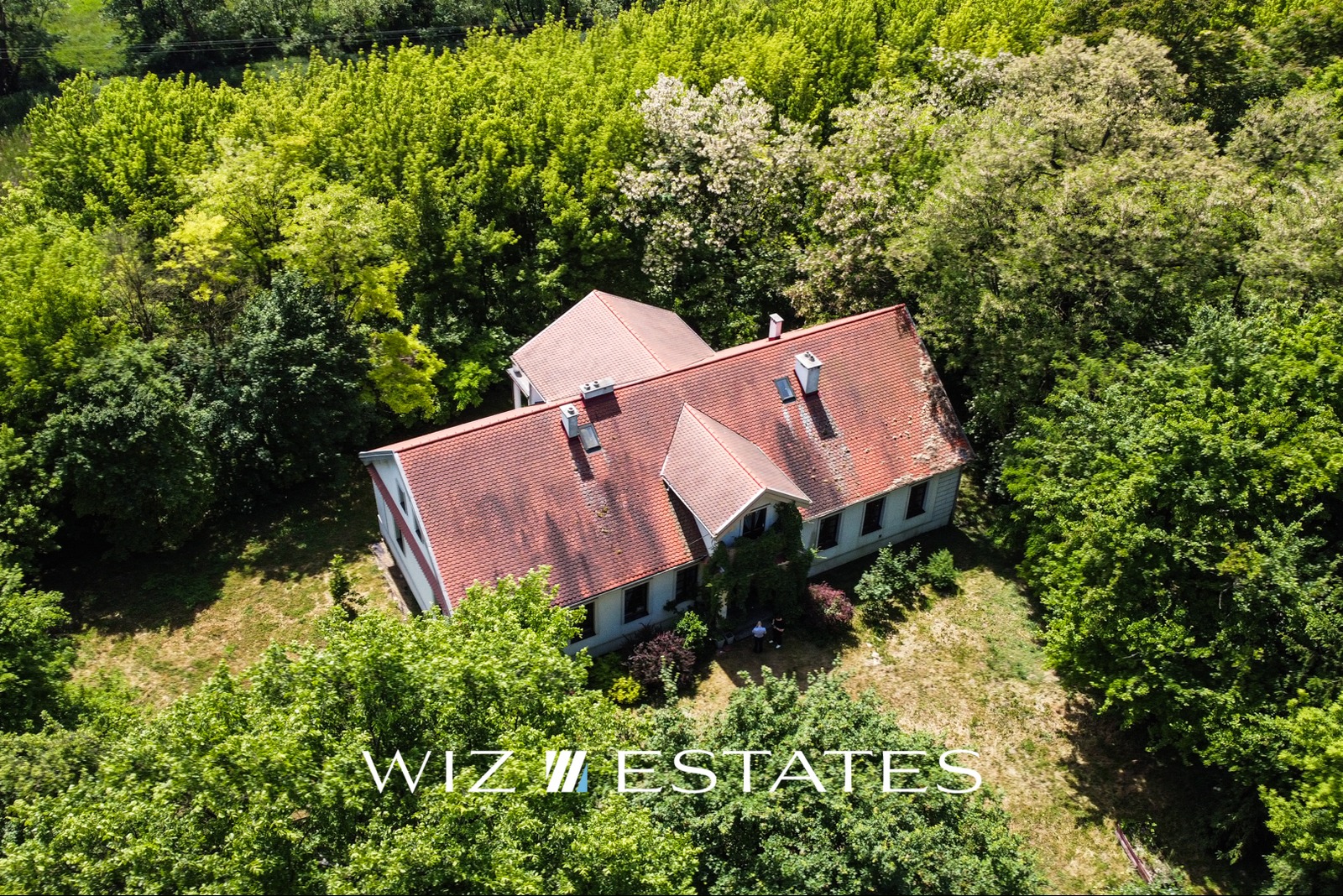 Dom z czerwonym dachem otoczony zielenią drzew, widok z lotu ptaka, logo WIZ Estates w dolnej części kadru.