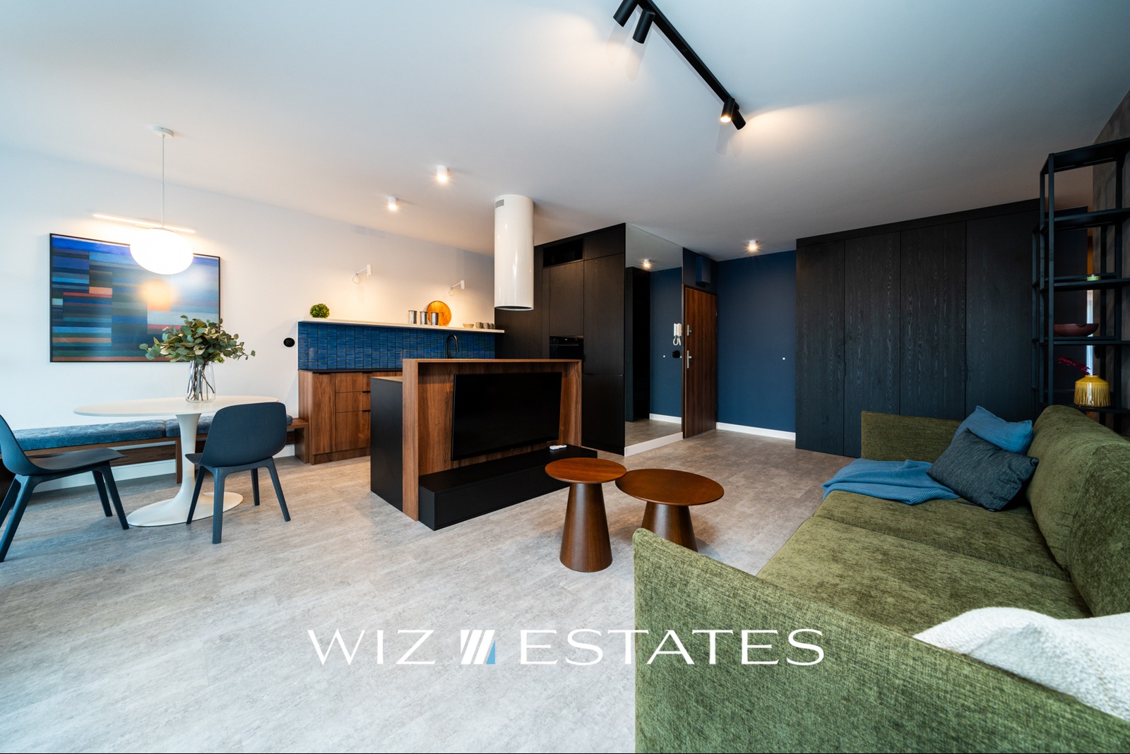 Jasne wnętrze mieszkania z aneksem kuchennym, okrągłym stołem z krzesłami i zieloną sofą, oświetlone punktowymi lampami.