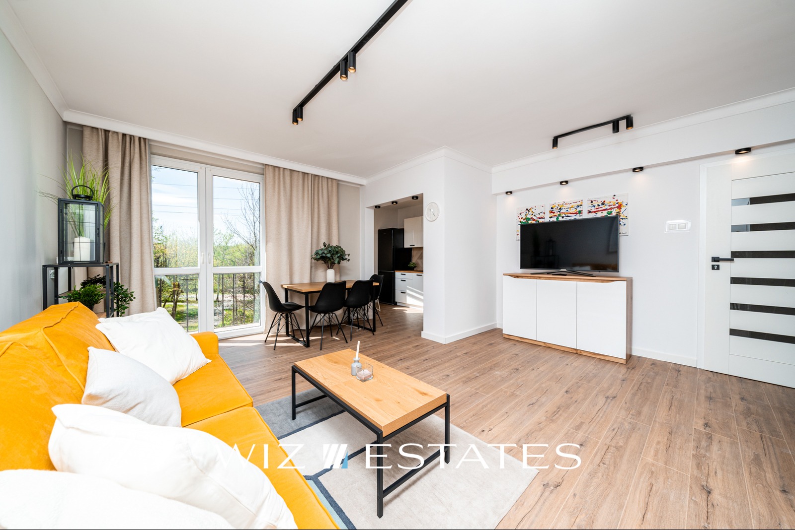 Jasne wnętrze mieszkania z żółtą sofą, drewnianym stolikiem kawowym i stołem jadalnym w tle, oświetlone nowoczesnymi lampami szynowymi.