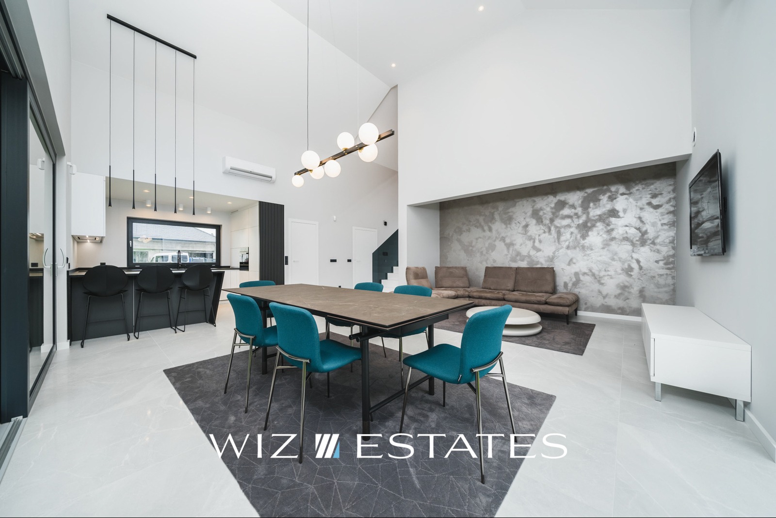 Jasne, przestronne wnętrze domu z wysokim sufitem, obejmujące otwartą kuchnię z czarnym barkiem, jadalnię z turkusowymi krzesłami, salon z szarą ścianą i brązową sofą, oraz białą podłogę z szarym...