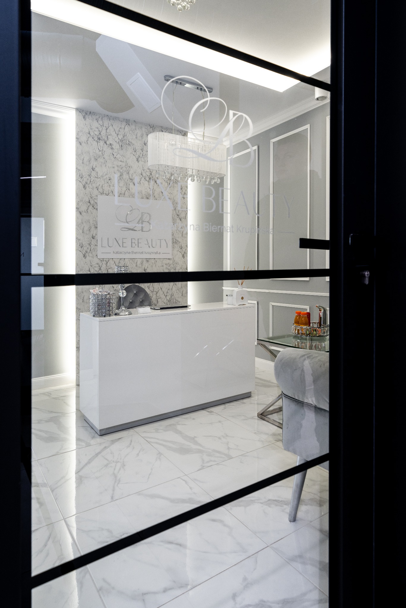 Nowoczesne wnętrze salonu kosmetycznego Luxe Beauty z białą ladą recepcyjną, marmurową ścianą i designerskim oświetleniem widziane przez przeszklone drzwi.