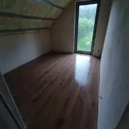 Poddasze po ociepleniu pianą i położeniu drewnopodobnej podłogi, widoczne metalowe profile sufitowe i okno dachowe z widokiem na zieleń.