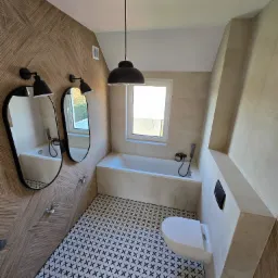 Nowoczesna łazienka z drewnopodobnymi płytkami na ścianie, geometryczną posadzką, owalnymi lustrami z lampkami i podwieszaną toaletą.