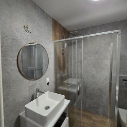 REMONTEX - wykończenia wnętrz RAFAŁ MARCZUK - Nowoczesna łazienka z szarymi płytkami imitującymi kamień, okrągłym lustrem w złotej ramie, białą umywalką nablatową na szafce i kabiną prysznicową ze szklanymi drzwiami, widoczne przewody...