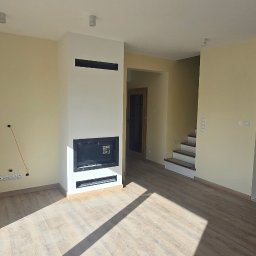 REMONTEX - wykończenia wnętrz RAFAŁ MARCZUK - Jasne wnętrze domu po remoncie: kremowe ściany, drewniana podłoga i nowoczesny kominek. Widoczne schody i okno. Częściowo wykończona podłoga z płytek.