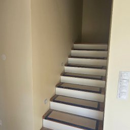 REMONTEX - wykończenia wnętrz RAFAŁ MARCZUK - Schody wewnętrzne po renowacji, z drewnianymi stopniami zabezpieczonymi taśmą malarską i białymi podstopnicami. Ściany w kolorze beżowym, oświetlenie punktowe.