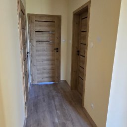 REMONTEX - wykończenia wnętrz RAFAŁ MARCZUK - Korytarz po remoncie: panele podłogowe imitujące drewno, dwoje drzwi z drewnianą okleiną i czarnymi klamkami, ściany w odcieniach beżu.