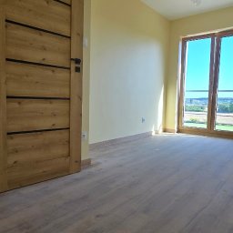 REMONTEX - wykończenia wnętrz RAFAŁ MARCZUK - Jasne, puste wnętrze po remoncie: drewniane drzwi z czarnymi akcentami, panele podłogowe w odcieniach brązu, ściany w odcieniach żółci, okno z widokiem na zieleń.