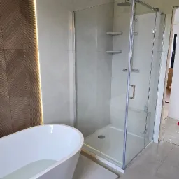 Nowoczesna łazienka z wolnostojącą wanną wypełnioną wodą i szklaną kabiną prysznicową. Brązowa ściana z oświetleniem LED. Szare płytki podłogowe.