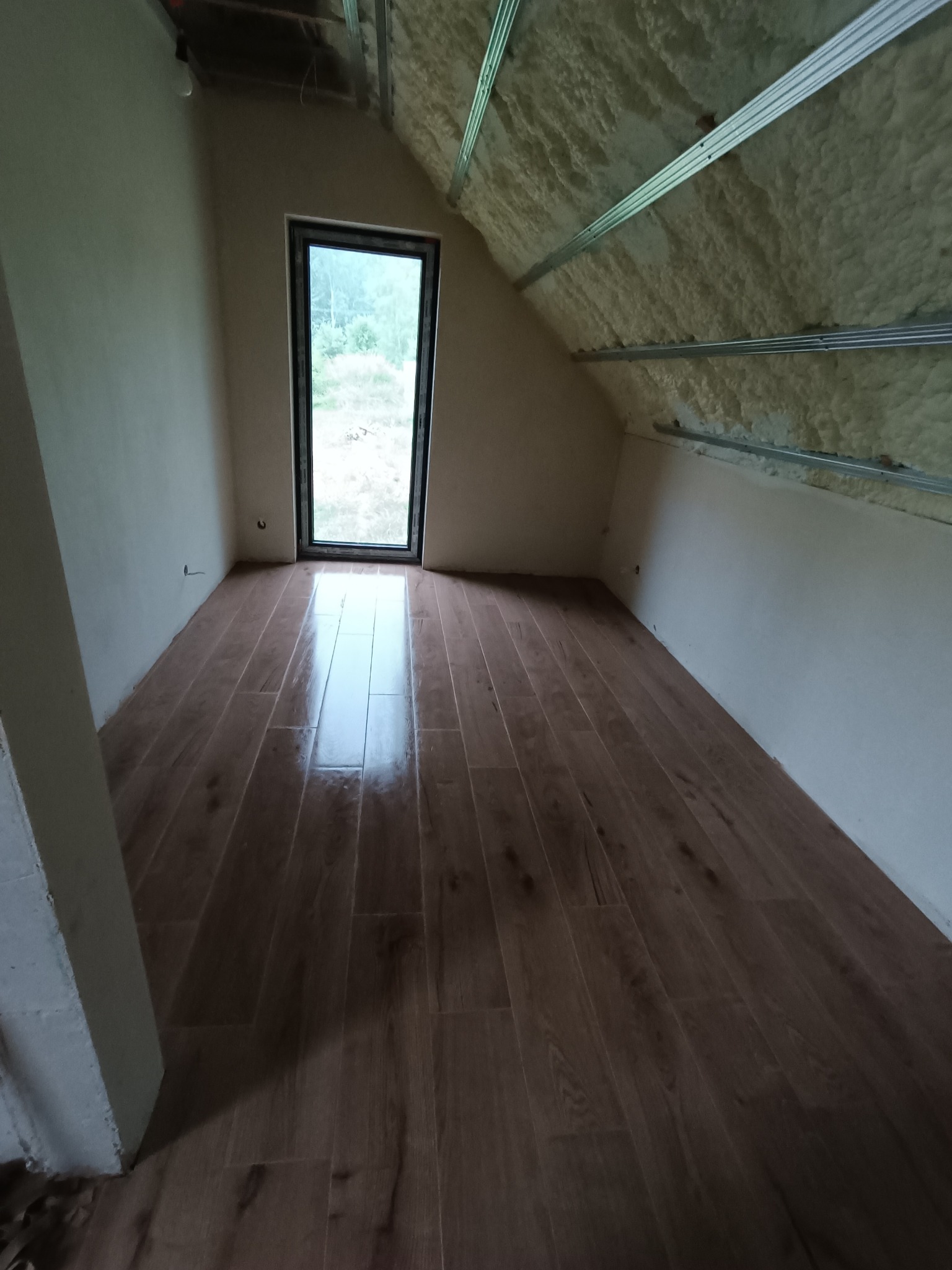 Poddasze po ociepleniu pianą, z zamontowaną konstrukcją pod płyty gipsowe i ułożoną podłogą z paneli imitujących drewno, widok na okno i fragment ściany.