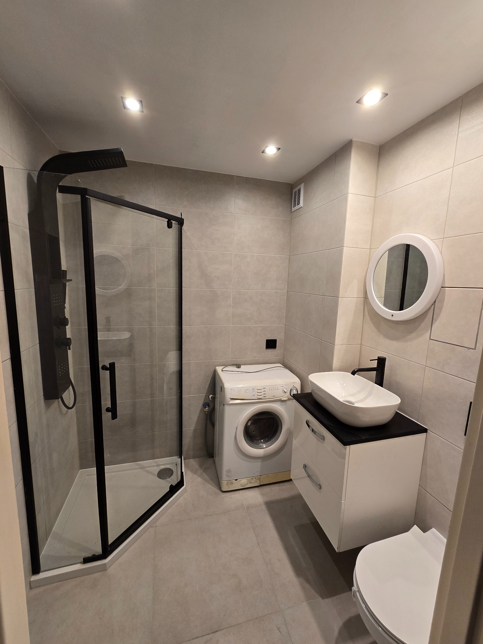 Nowoczesna łazienka z kabiną prysznicową typu walk-in z czarnymi ramami, białą umywalką nablatową na szafce z szufladami, okrągłym lustrem z podświetleniem LED, pralką i płytkami w odcieniach...