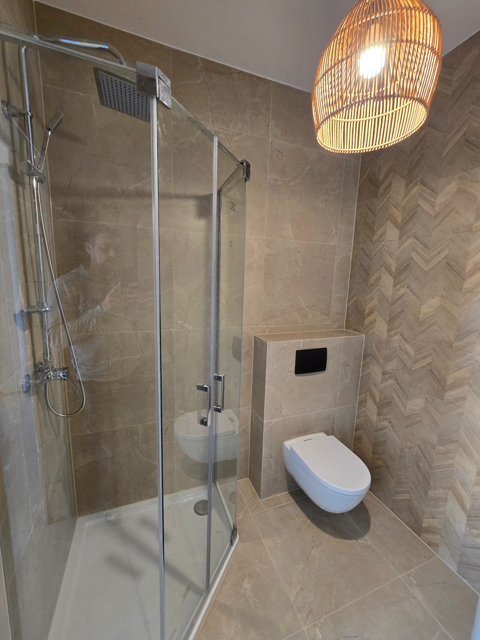 Nowoczesna łazienka z kabiną prysznicową typu walk-in, podwieszaną toaletą i dekoracyjną ścianą w jodełkę, oświetlona lampą z wikliny.