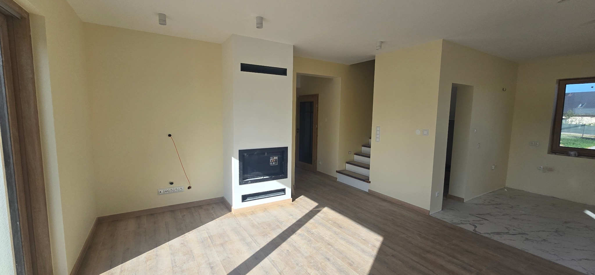 Jasne wnętrze domu po remoncie: kremowe ściany, drewniana podłoga i nowoczesny kominek. Widoczne schody i okno. Częściowo wykończona podłoga z płytek.