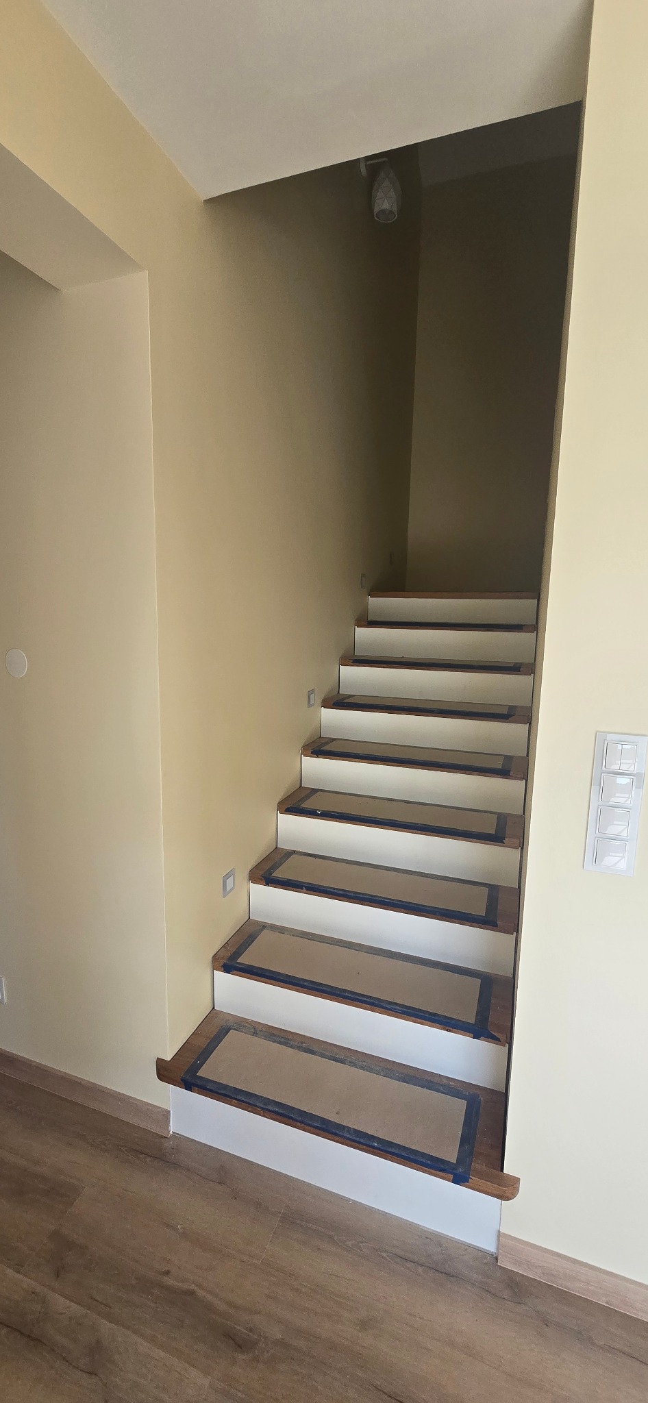 Schody wewnętrzne po renowacji, z drewnianymi stopniami zabezpieczonymi taśmą malarską i białymi podstopnicami. Ściany w kolorze beżowym, oświetlenie punktowe.
