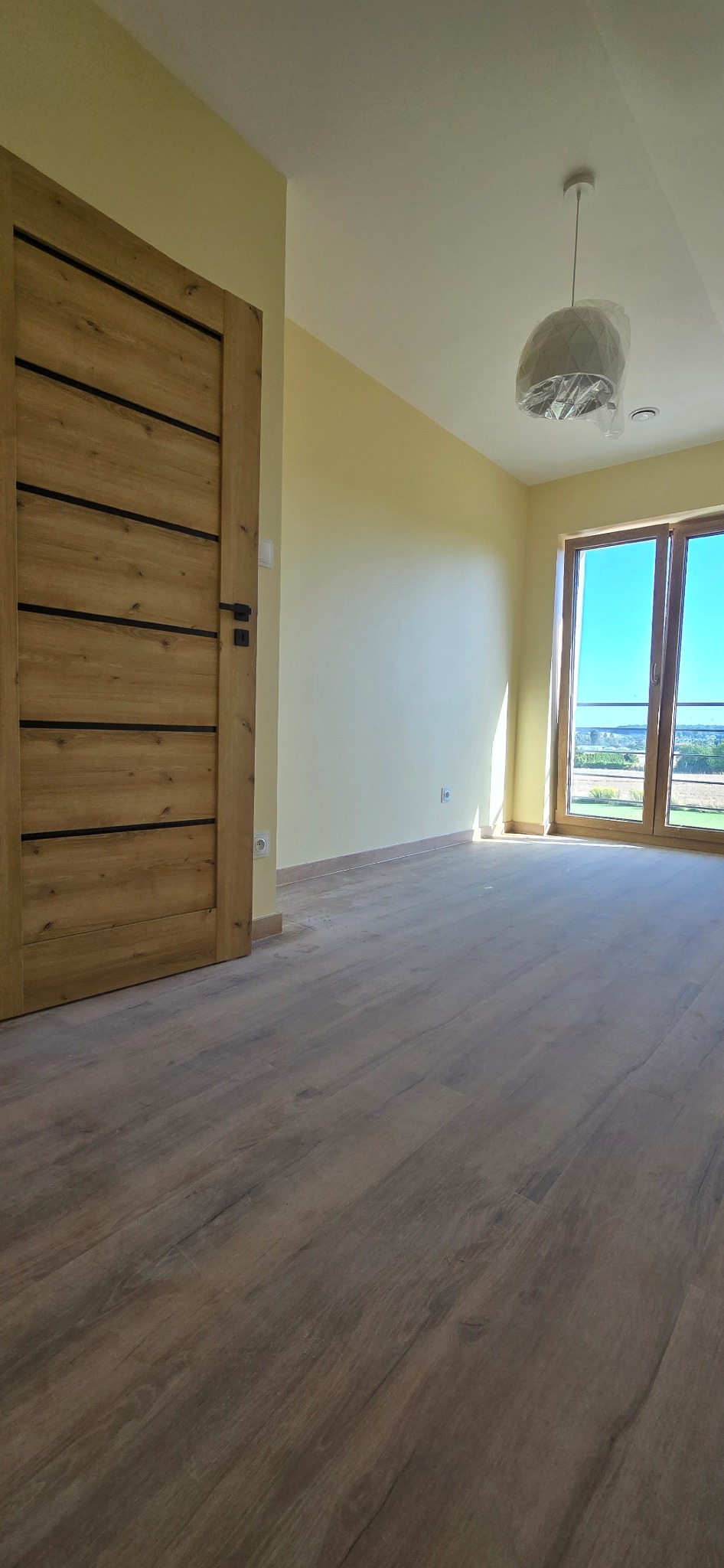 Jasne, puste wnętrze po remoncie: drewniane drzwi z czarnymi akcentami, panele podłogowe w odcieniach brązu, ściany w odcieniach żółci, okno z widokiem na zieleń.