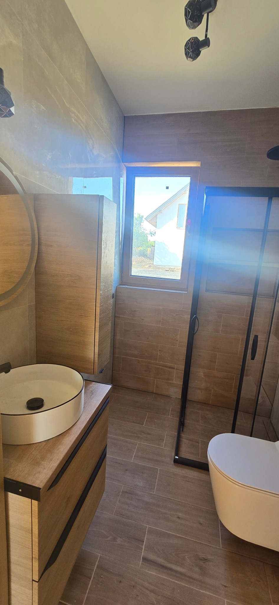 Nowoczesna łazienka z drewnopodobnymi płytkami, kabiną prysznicową typu walk-in i umywalką nablatową. Widoczne okno z widokiem na dom.