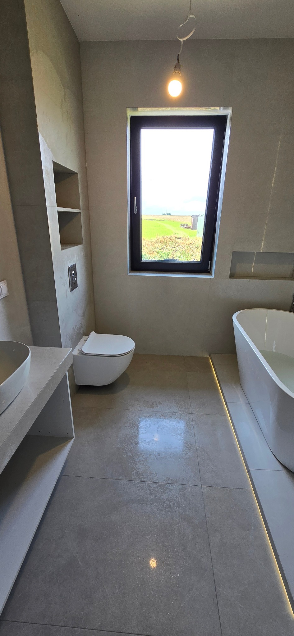 Nowoczesna łazienka w odcieniach szarości z oknem na zielony krajobraz. Minimalistyczna armatura, podświetlana podłoga i surowe ściany tworzą spójny design.