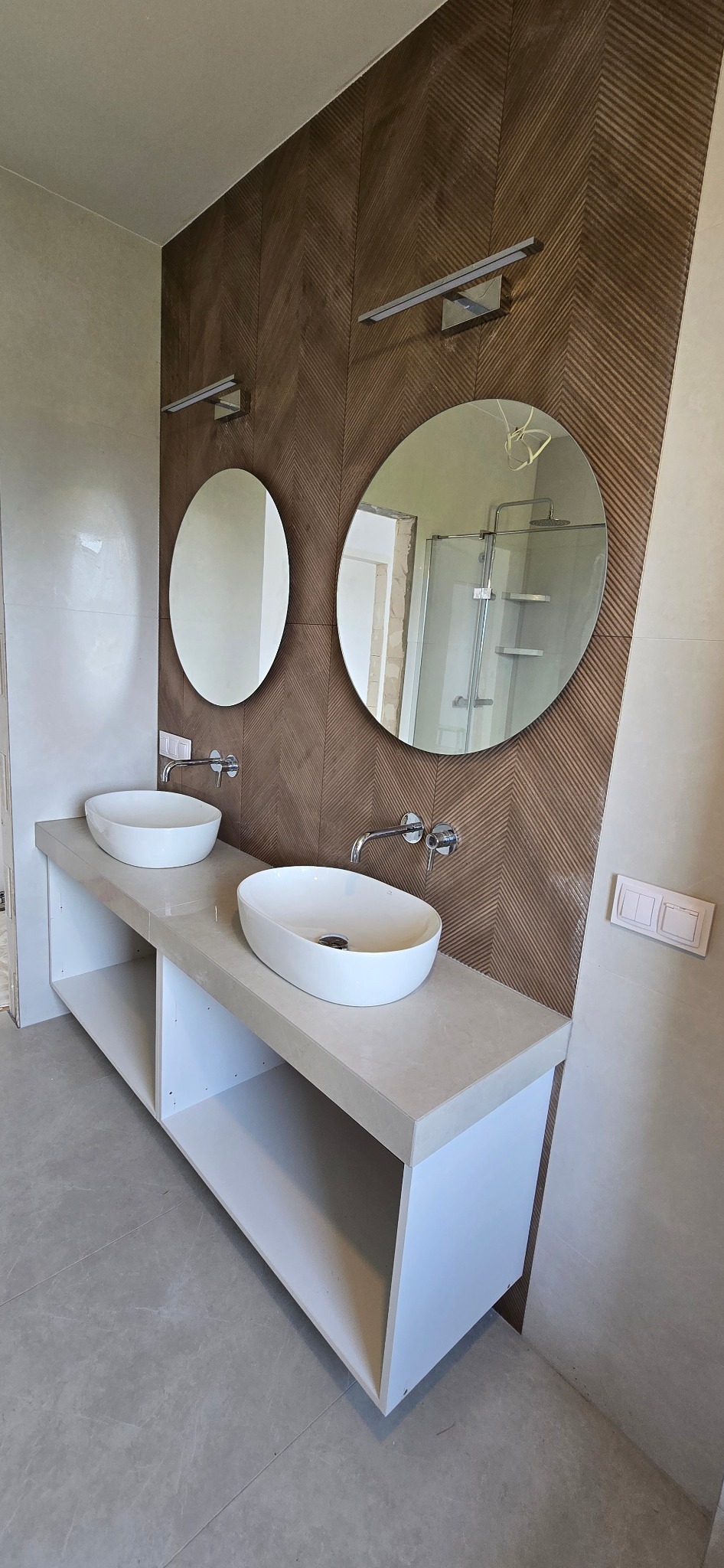 Nowoczesna łazienka z dwoma owalnymi umywalkami na blacie z półkami. Na ścianie ozdobna, brązowa okładzina w jodełkę i okrągłe lustra z oświetleniem.