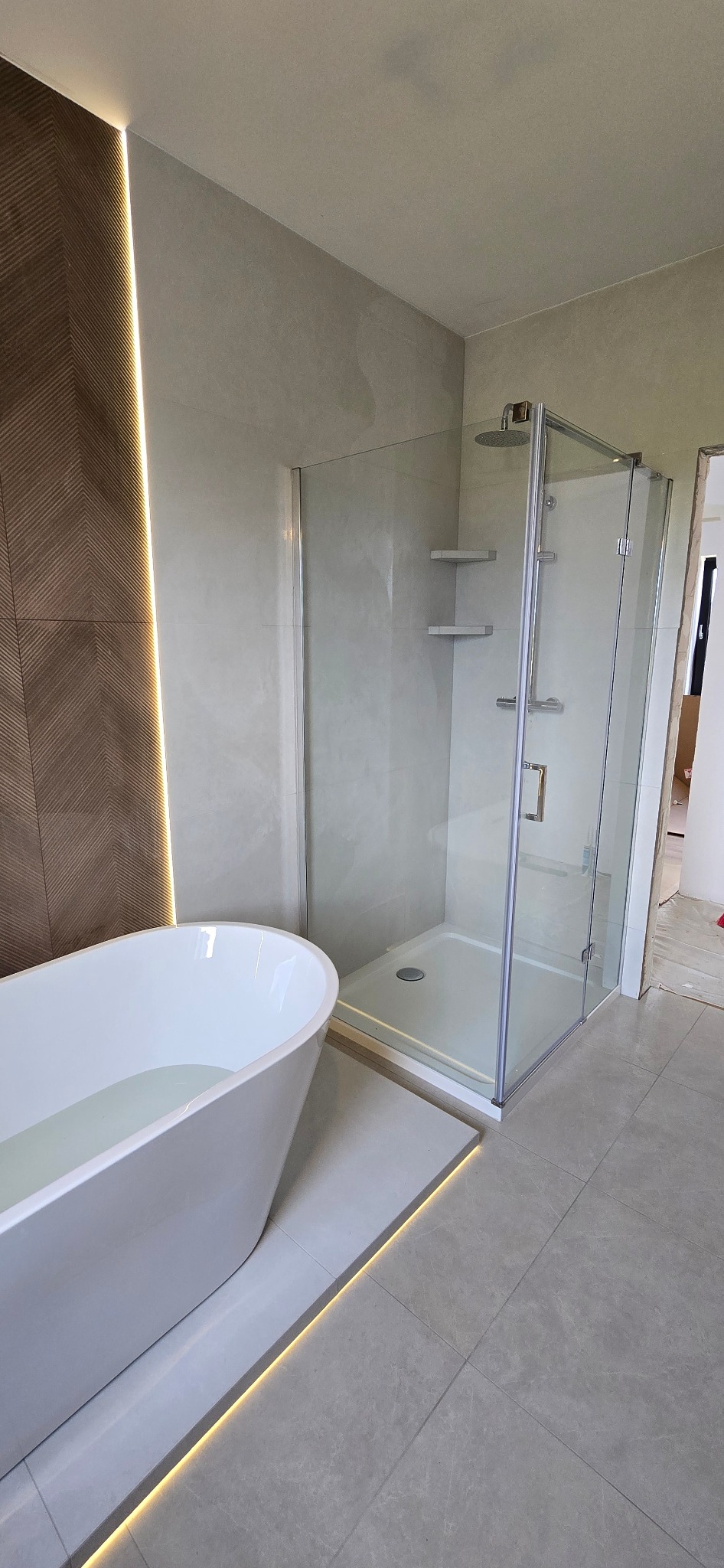 Nowoczesna łazienka z wolnostojącą wanną wypełnioną wodą i szklaną kabiną prysznicową. Brązowa ściana z oświetleniem LED. Szare płytki podłogowe.