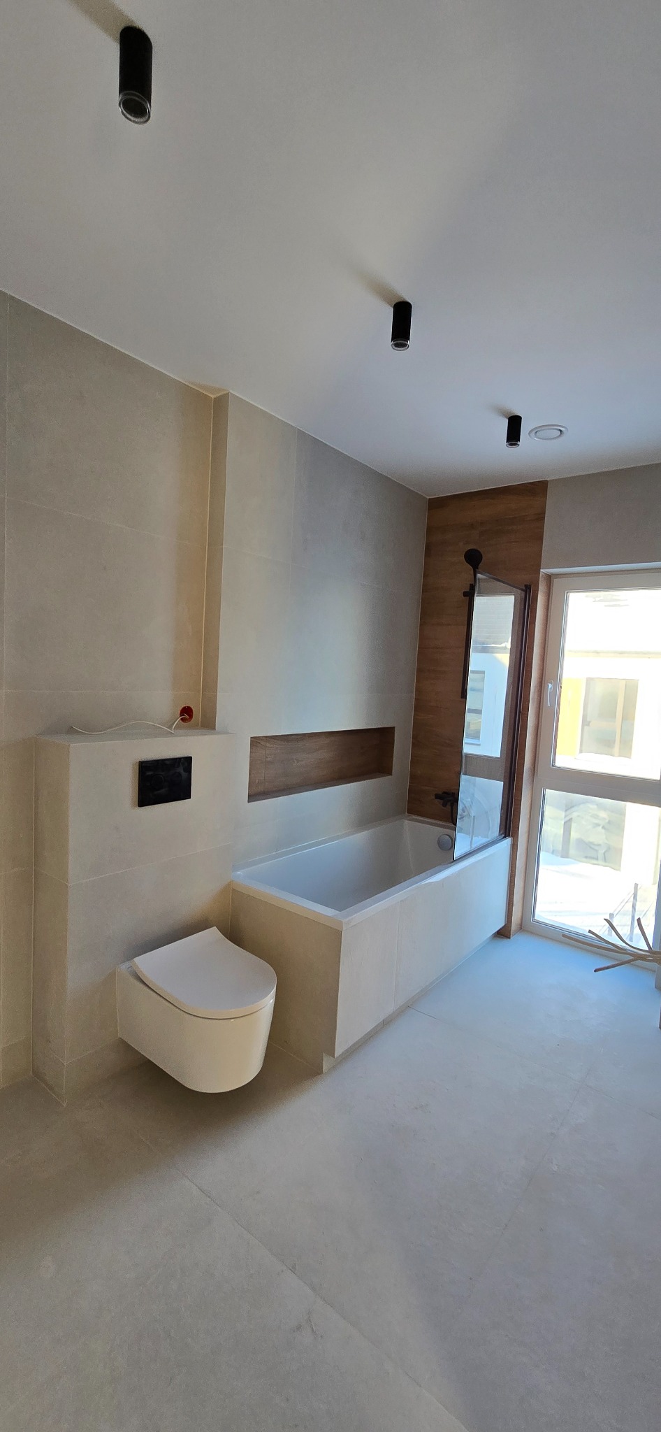 Nowoczesna łazienka z beżowymi płytkami, podwieszaną toaletą i wanną z parawanem. Minimalistyczne oświetlenie punktowe i drewniane akcenty dekoracyjne.