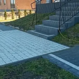 Ułożona kostka granitowa w odcieniach szarości tworząca chodnik i schody przed domem z czarną balustradą, widoczny trawnik i fragment elewacji budynku