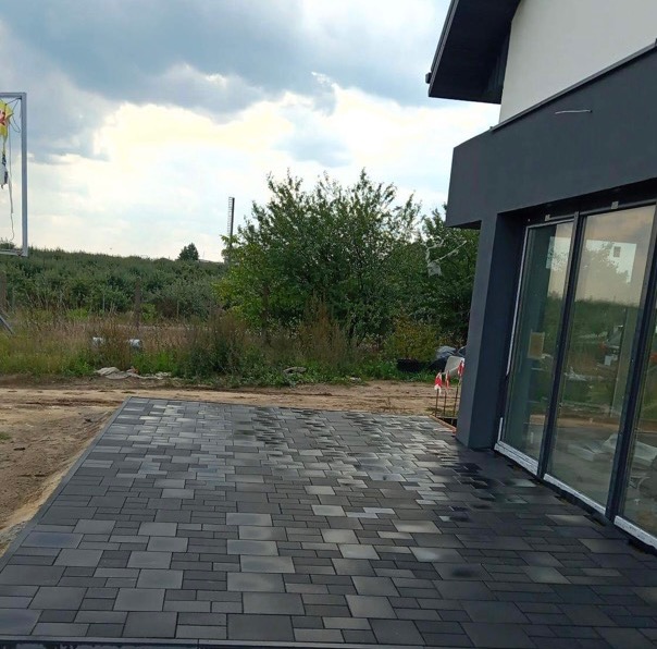 Ułożona, szara kostka brukowa na posesji przed nowoczesnym domem z przesuwnymi drzwiami tarasowymi, widoczne tło z zielenią i pochmurne niebo.